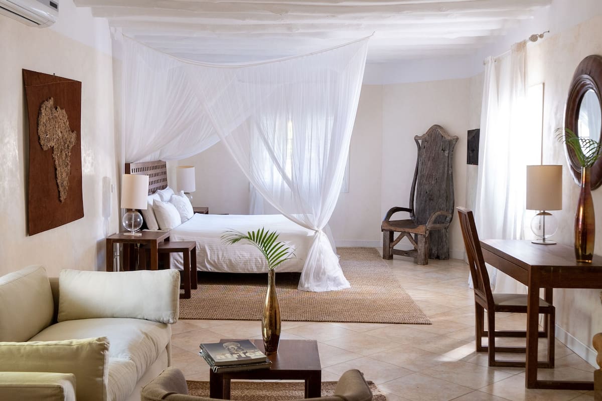 A bedroom with a canopy bed, a sitting area with a table and chairs, and a bedside table with a lamp and a plant.