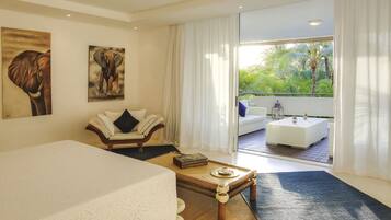 A hotel room with a bed, a wooden coffee table, a white sofa, and a balcony with a view of greenery.