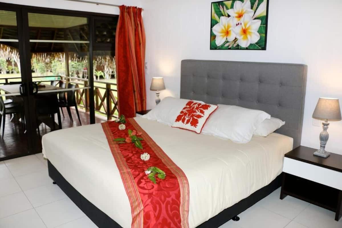 A hotel room with a bed, a nightstand, a lamp, a painting of flowers, and a view of an outdoor area with tables and chairs.