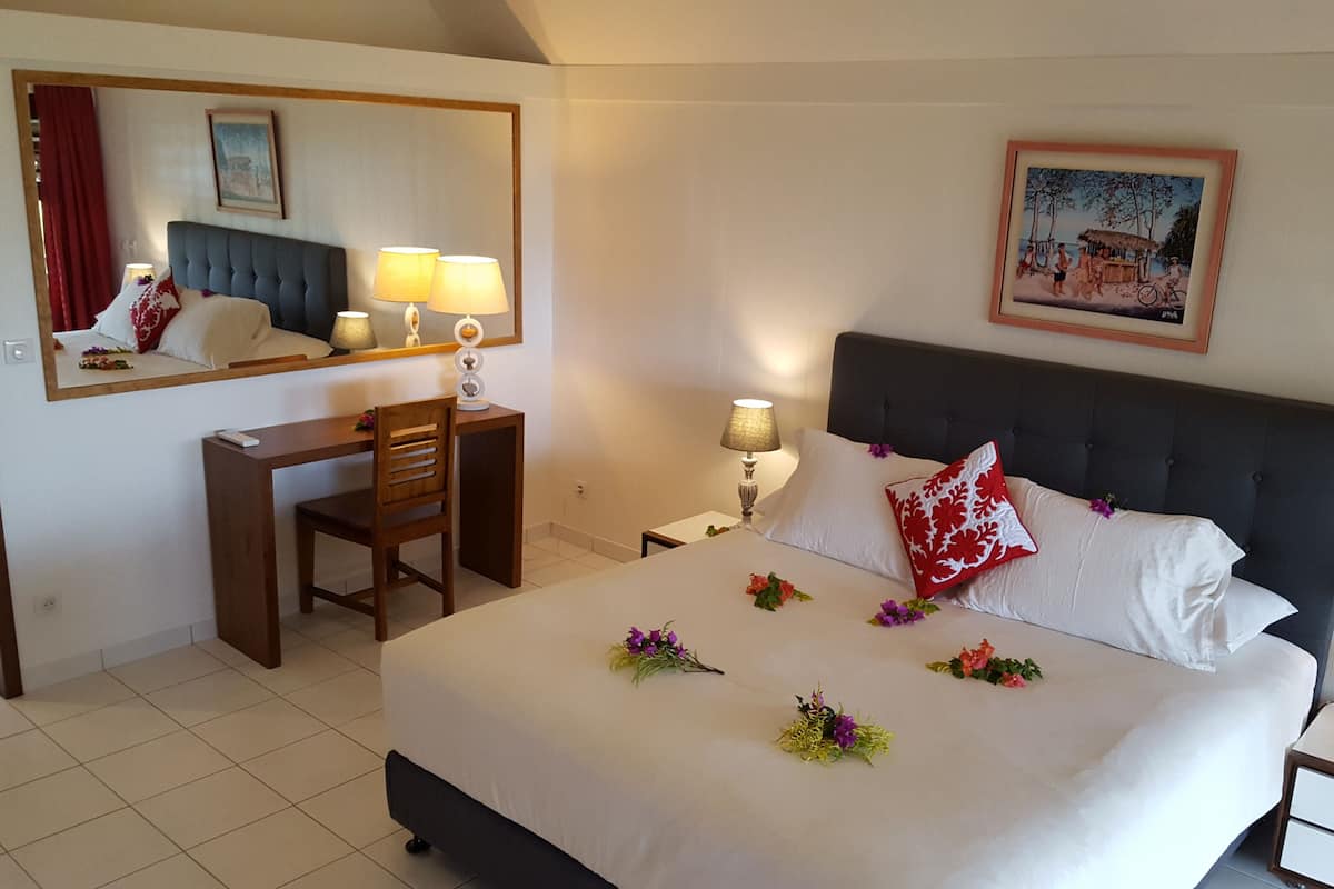 A hotel room with a bed, a desk, a chair, a nightstand, a lamp, a mirror, and a framed picture on the wall.