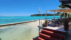 Une terrasse au bord de la plage, avec une eau turquoise et limpide, un escalier en bois menant à la plage, et des espaces de détente en plein air avec des parasols.