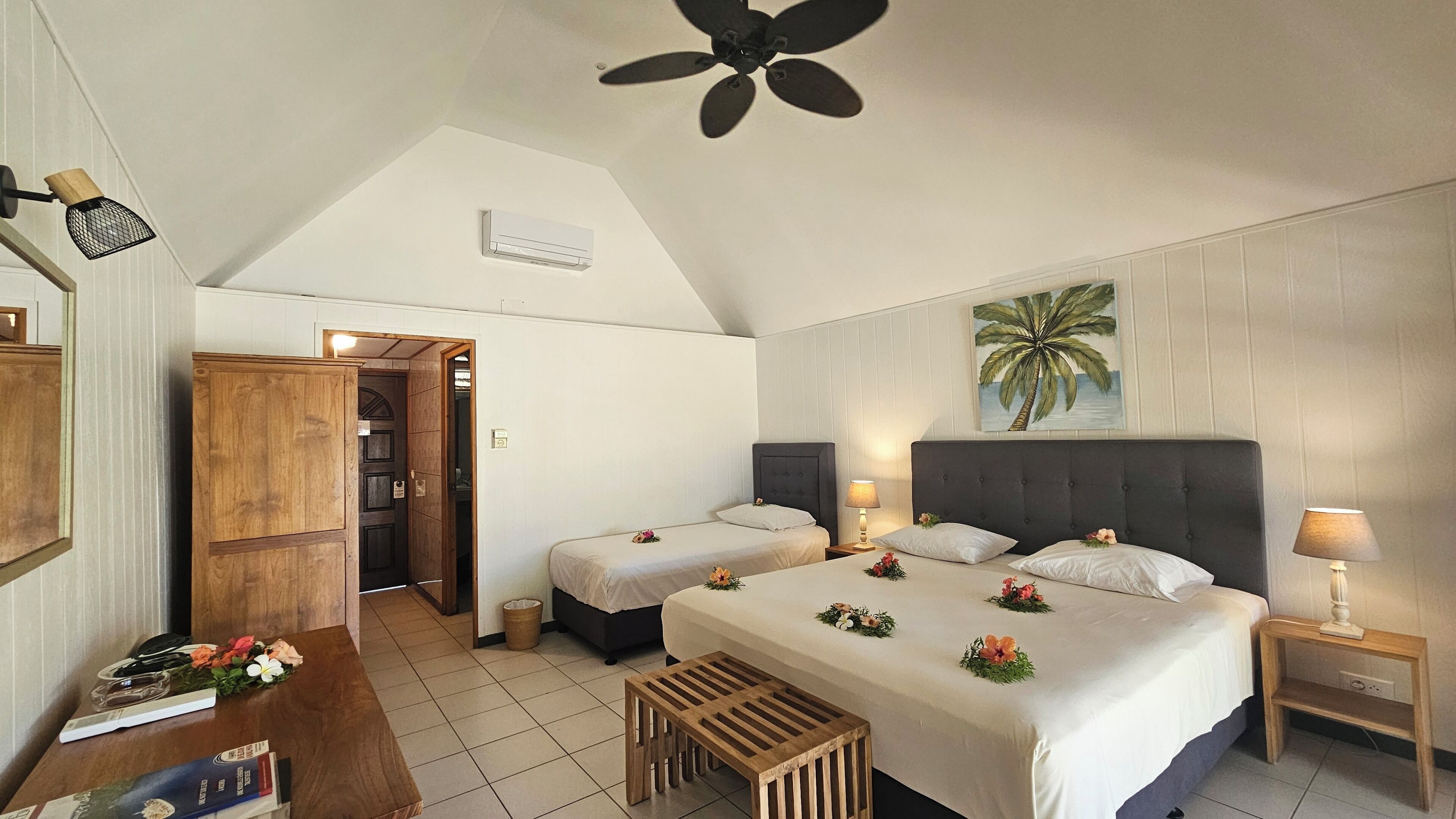 A bedroom with two beds, a ceiling fan, a painting of a palm tree, and a wooden wardrobe.