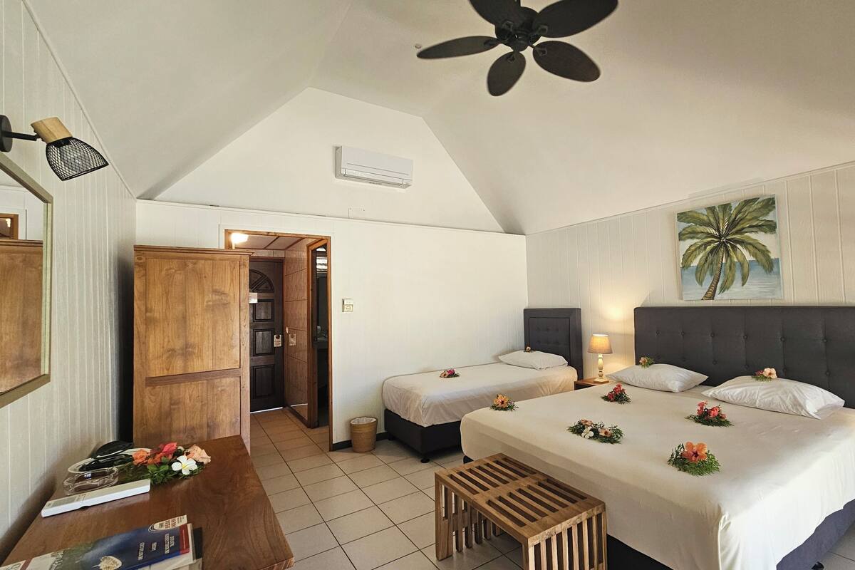 A bedroom with two beds, a ceiling fan, a painting of a palm tree, and a wooden wardrobe.