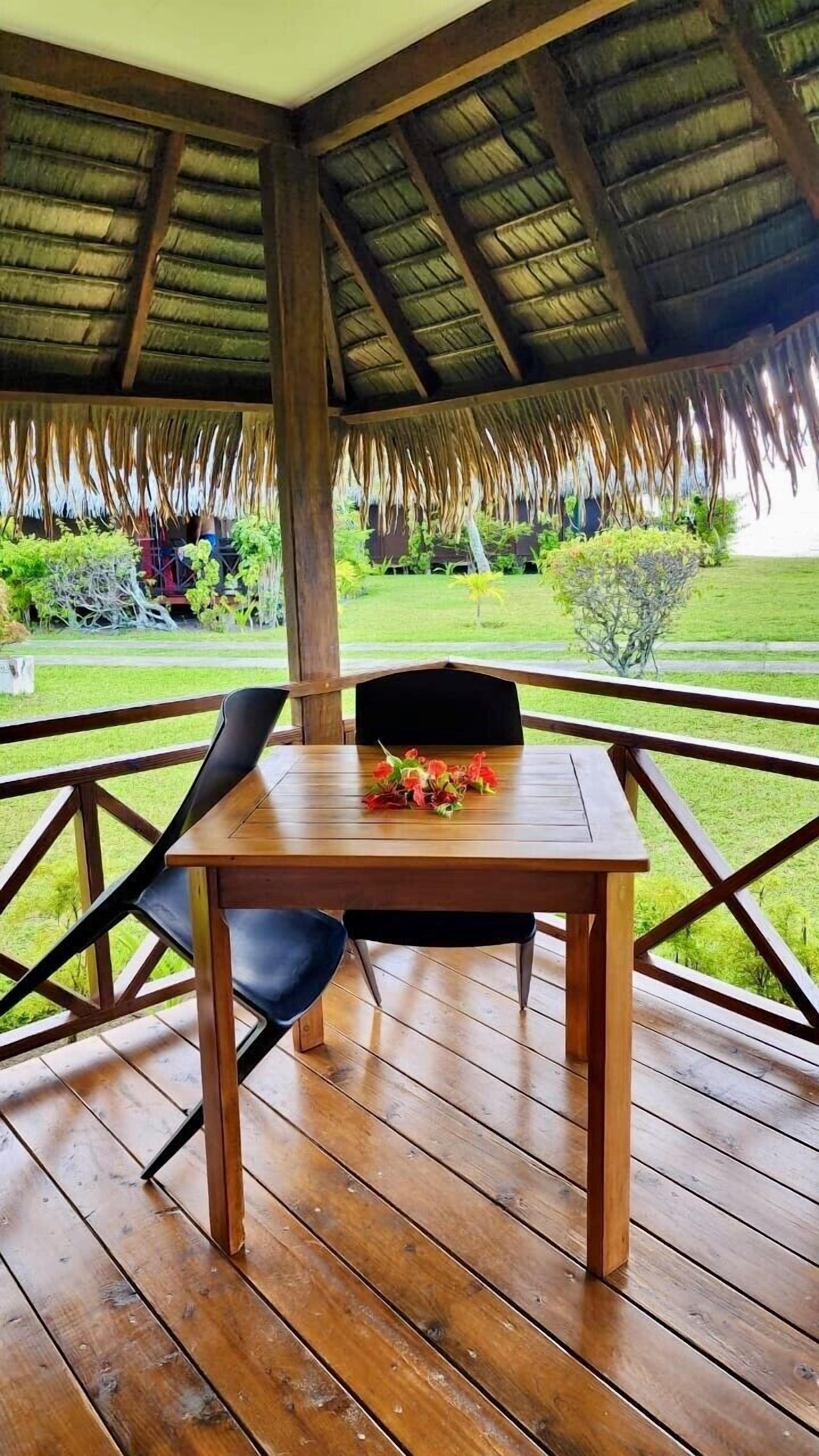 Une terrasse en bois avec une table et des chaises, un toit de chaume et une vue sur un jardin luxuriant.