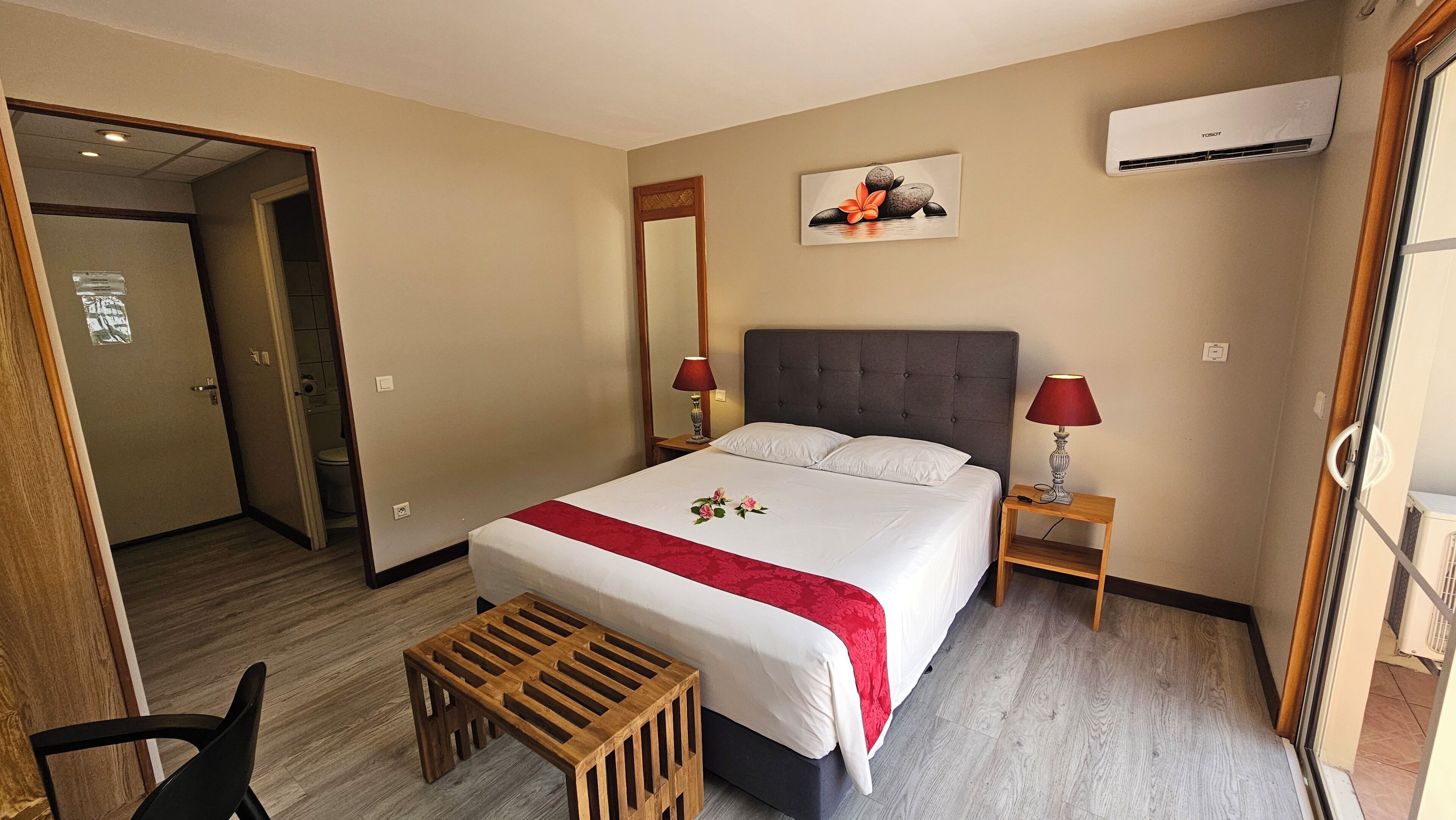 Une chambre d’hôtel comprenant un lit, des tables de chevet, un tabouret en bois, un miroir, un appareil de climatisation et une porte donnant sur une salle de bain.