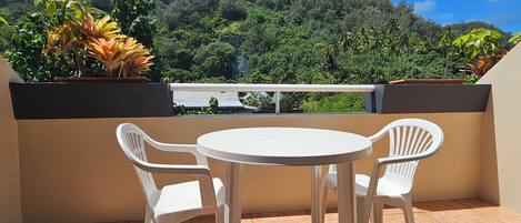 Un balcón con dos sillas de plástico blanco y una pequeña mesa redonda, con vista a un paisaje verde exuberante con árboles y una montaña al fondo.