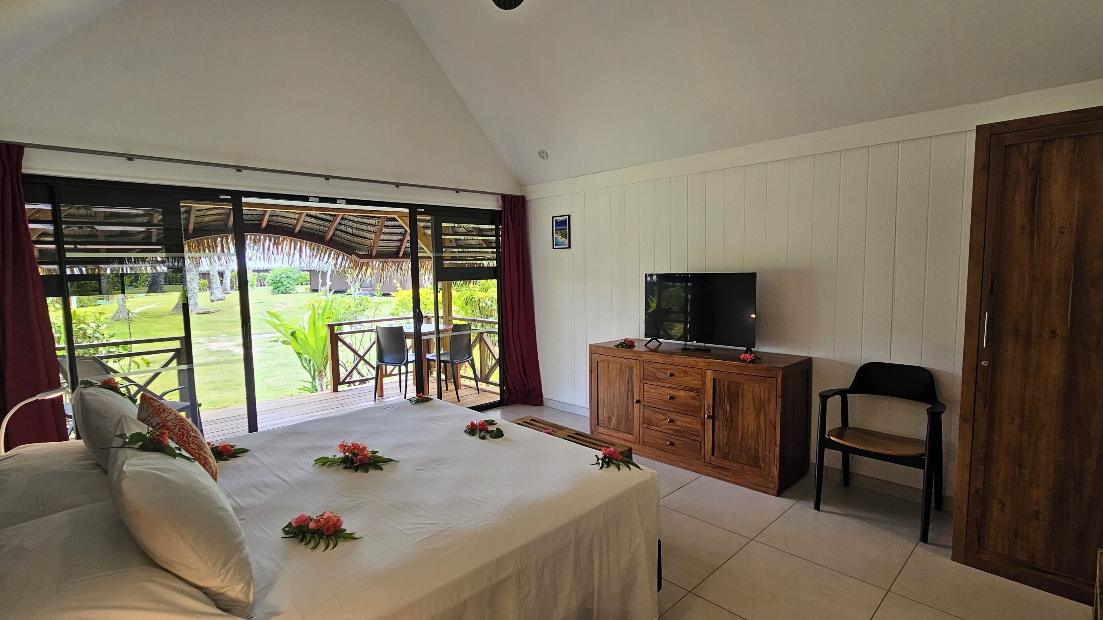 Een slaapkamer met een bed, een televisie op een houten kast, een zithoek met stoelen en een tafel, en uitzicht op een tuin door grote ramen.