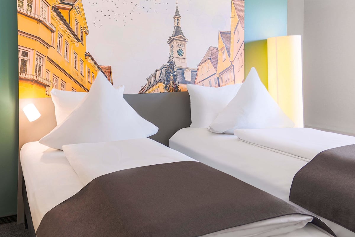Une chambre d’hôtel avec deux lits, du linge de lit blanc et une grande fresque murale représentant un paysage urbain.