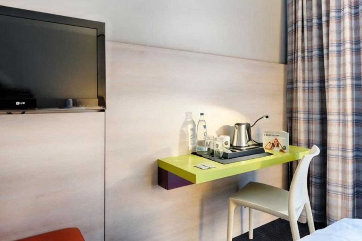 Habitación de hotel con una televisión de pantalla plana montada en la pared, un escritorio pequeño con una silla y una cama con cabecera blanca.