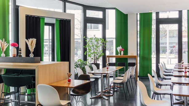 Un comedor moderno con varias mesas y sillas, ventanas amplias con cortinas verdes y plantas de interior.