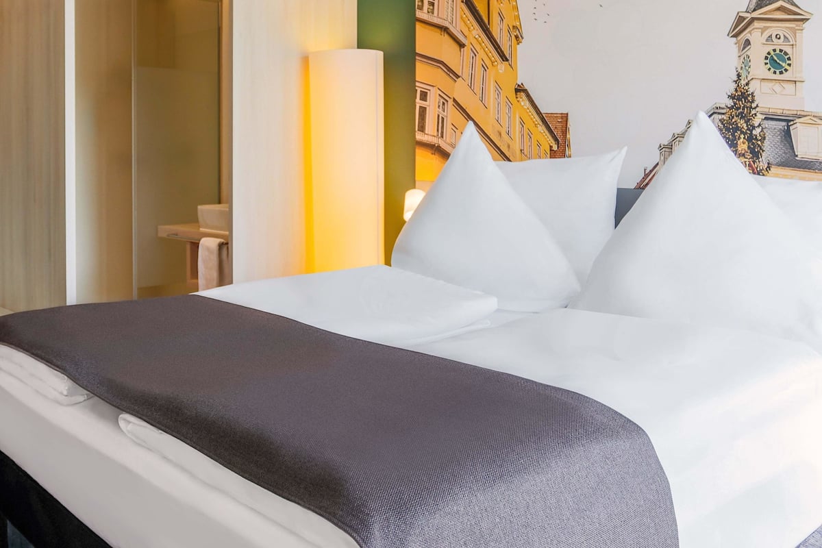 Une chambre d’hôtel avec un lit, des oreillers, une table de chevet et une fresque murale représentant un bâtiment.