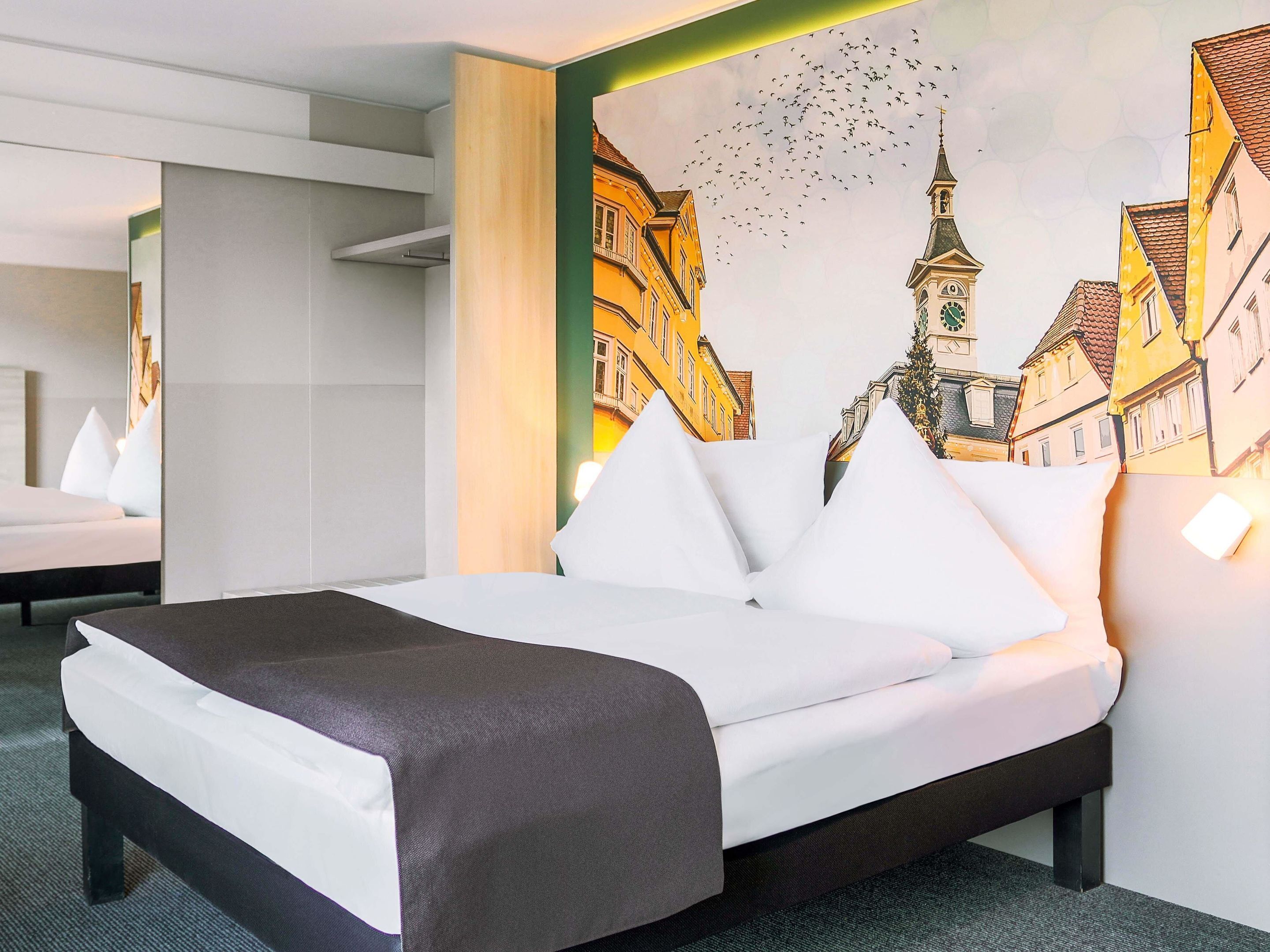 A hotel room with a bed, a large painting of a cityscape, and a mirror reflecting the room.