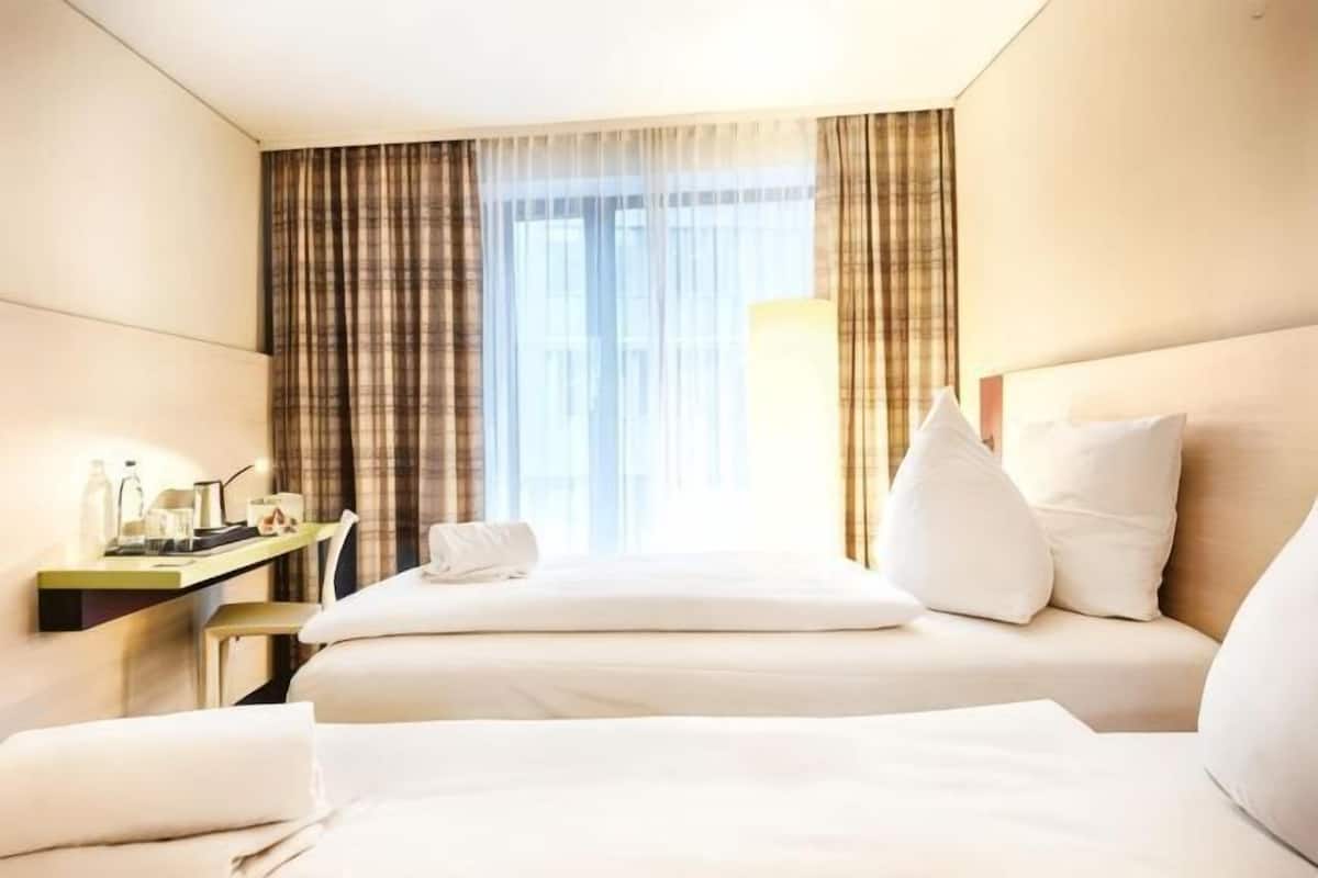 Une chambre d’hôtel avec deux lits, un bureau avec une lampe et une fenêtre avec des rideaux.