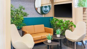Un área de descanso moderna con un sofá, dos sillones, una mesita de centro y varias plantas en macetas. Un espejo grande y un televisor de pantalla plana están montados en la pared.