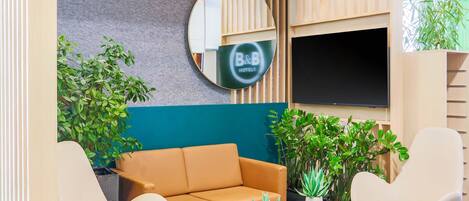 A modern lounge area with a sofa, two armchairs, a coffee table, and several potted plants. A large mirror and a flat-screen TV are mounted on the wall.