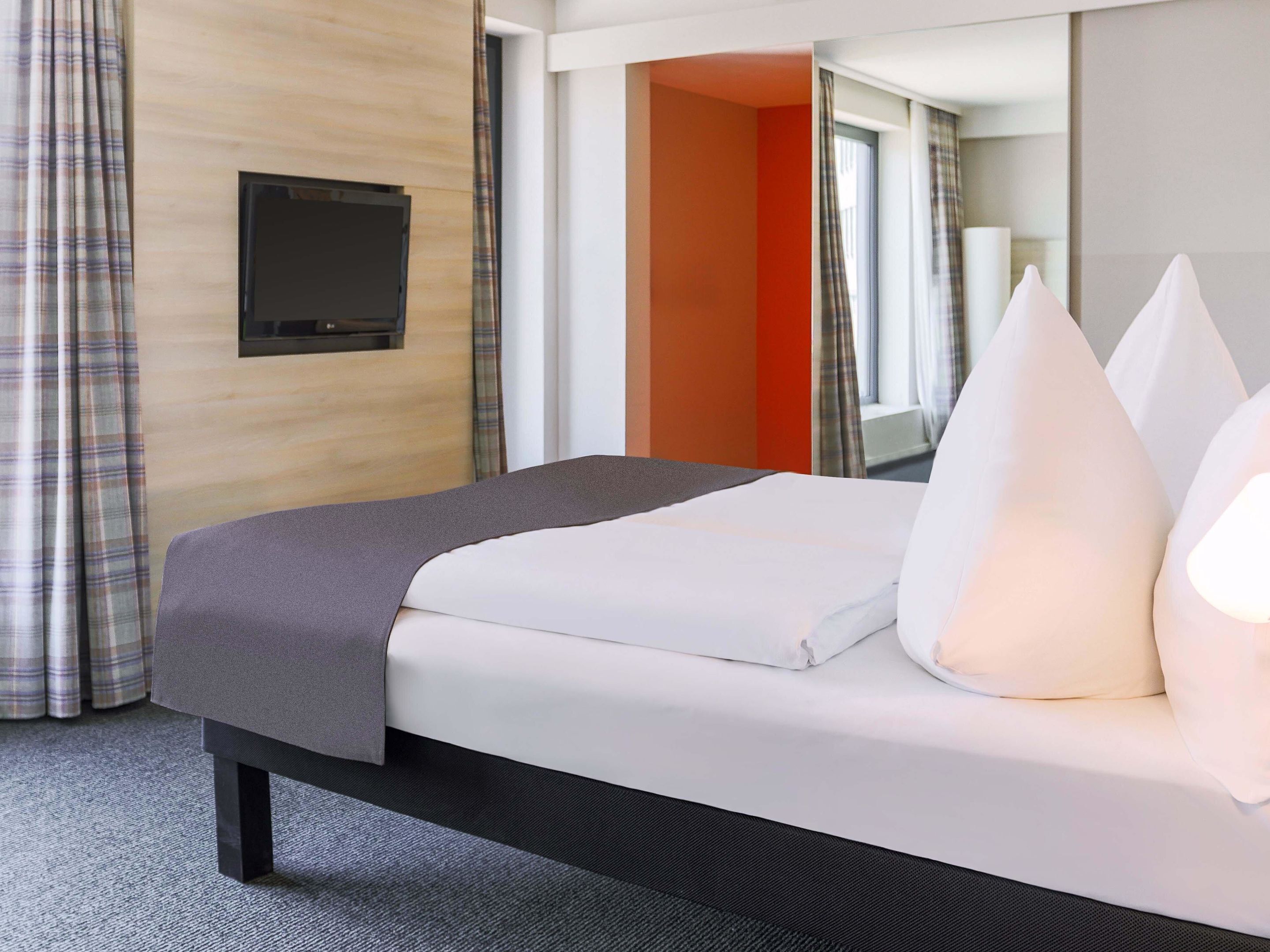 A hotel room with a bed, a flat-screen TV mounted on the wall, and a window with curtains.