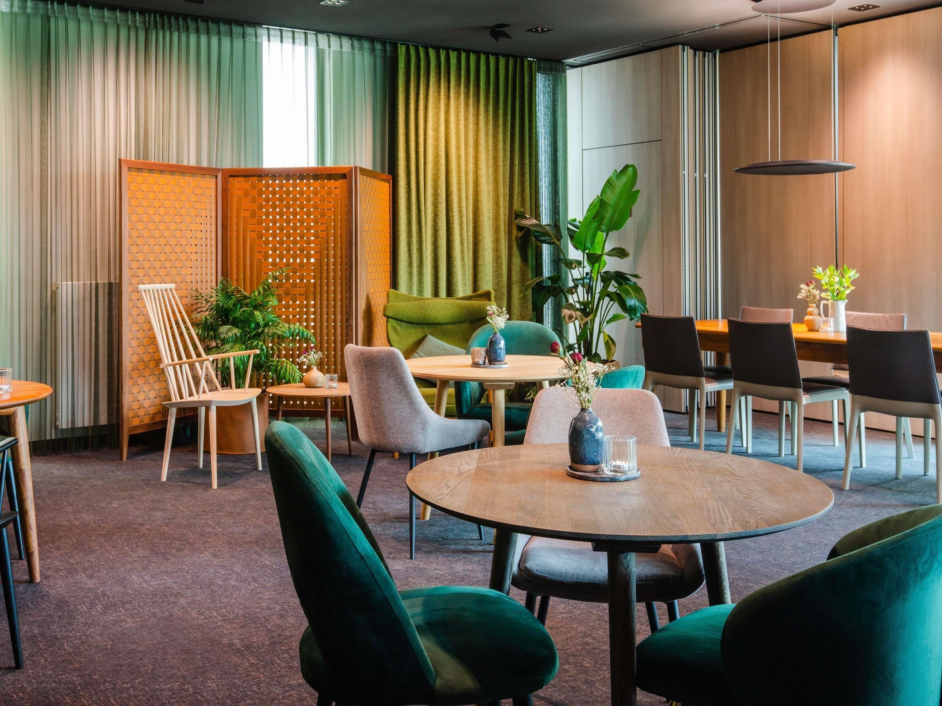 A modern dining area with round tables, green upholstered chairs, and a variety of plants.