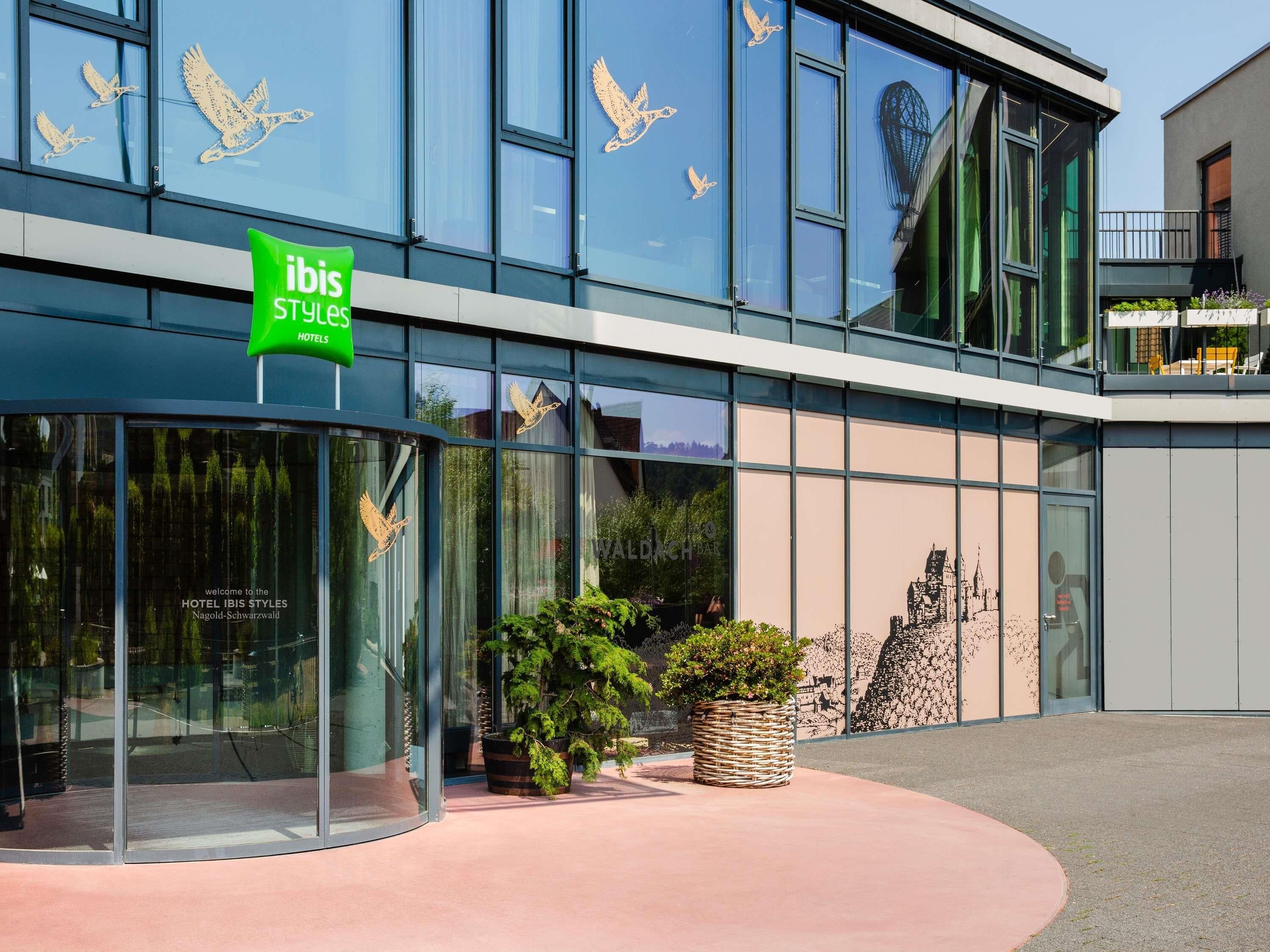 Un hotel moderno con grandes ventanales, un letrero verde que indica 'IBIS STYLES HOTELS' y una pared decorativa con motivos de aves.