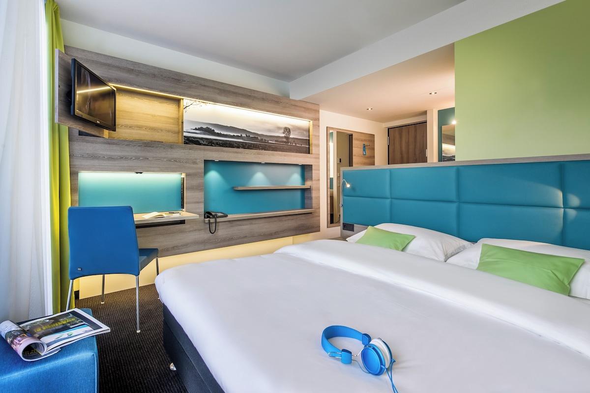 Una habitación de hotel moderna con una cama grande, un escritorio con computadora, un televisor en la pared y vistas al exterior.