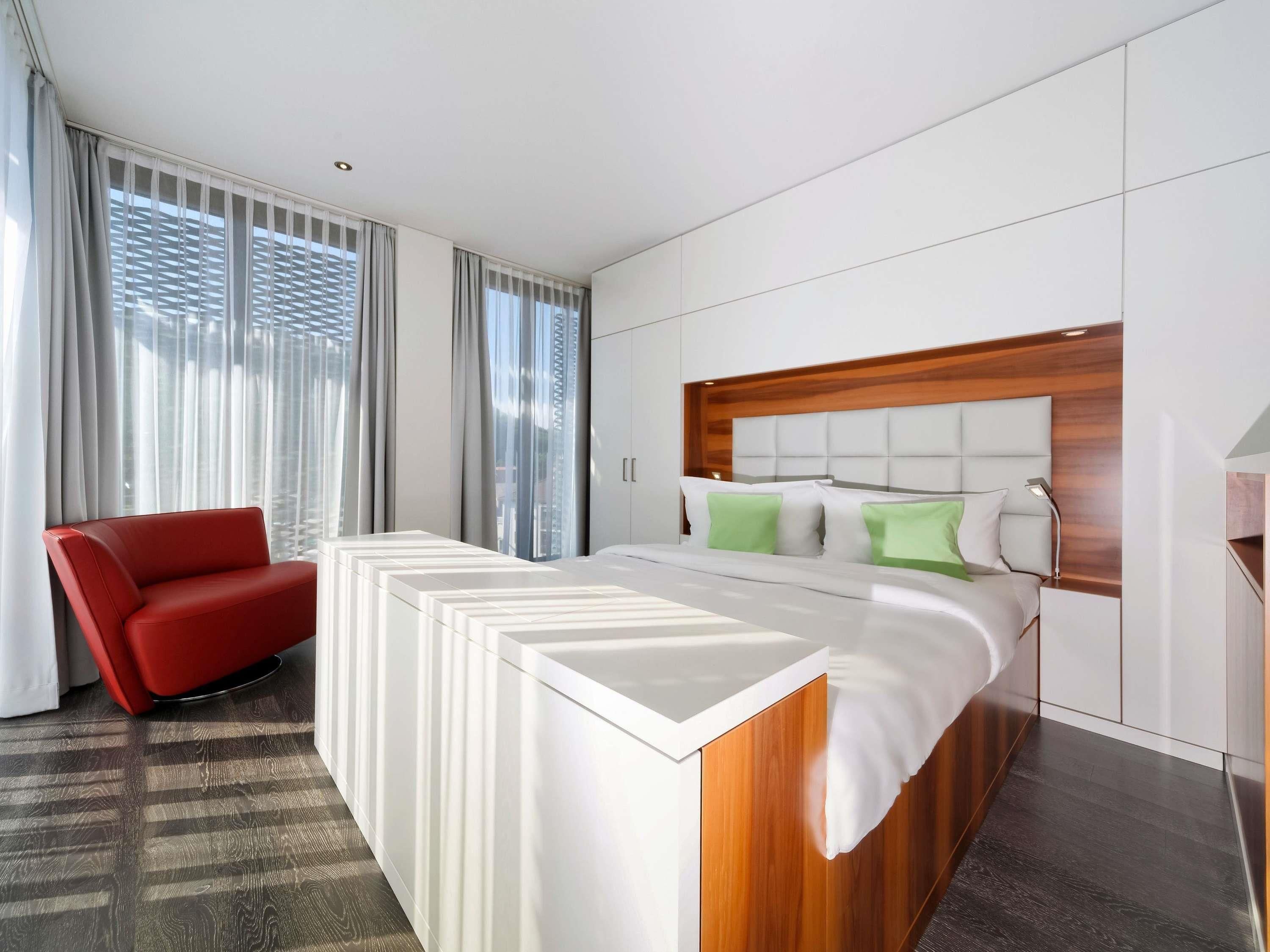 A modern hotel room with a large bed, a red chair, and a view through large windows.