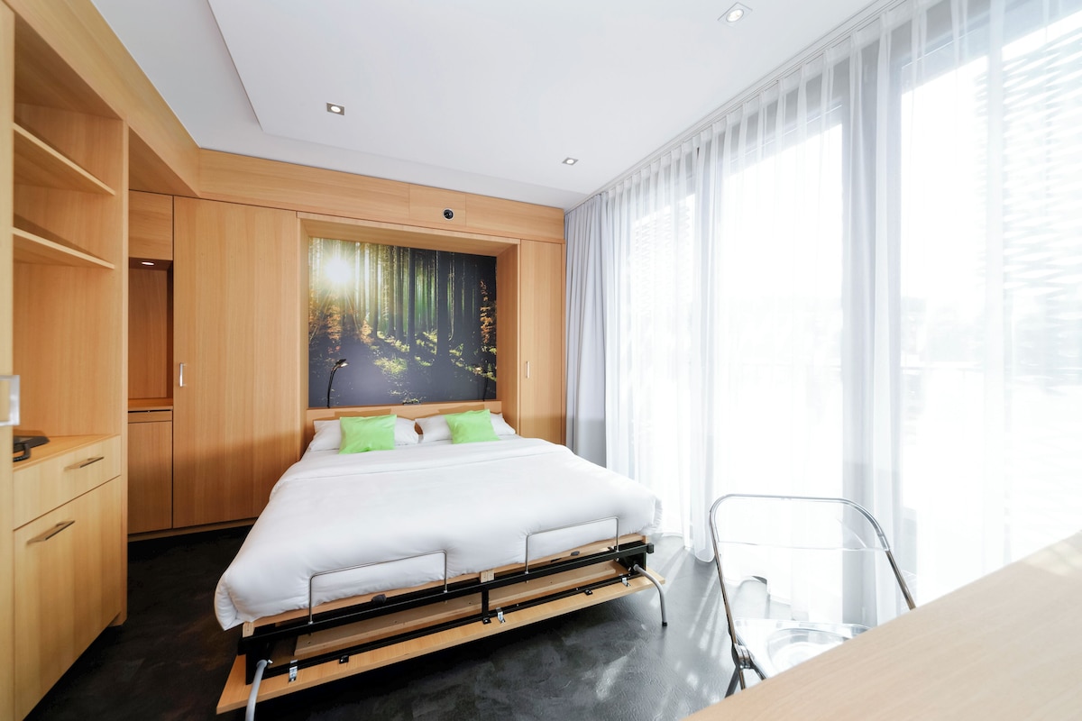 Una habitación de hotel moderna con una cama, armario de madera y un ventanal con cortinas translúcidas.