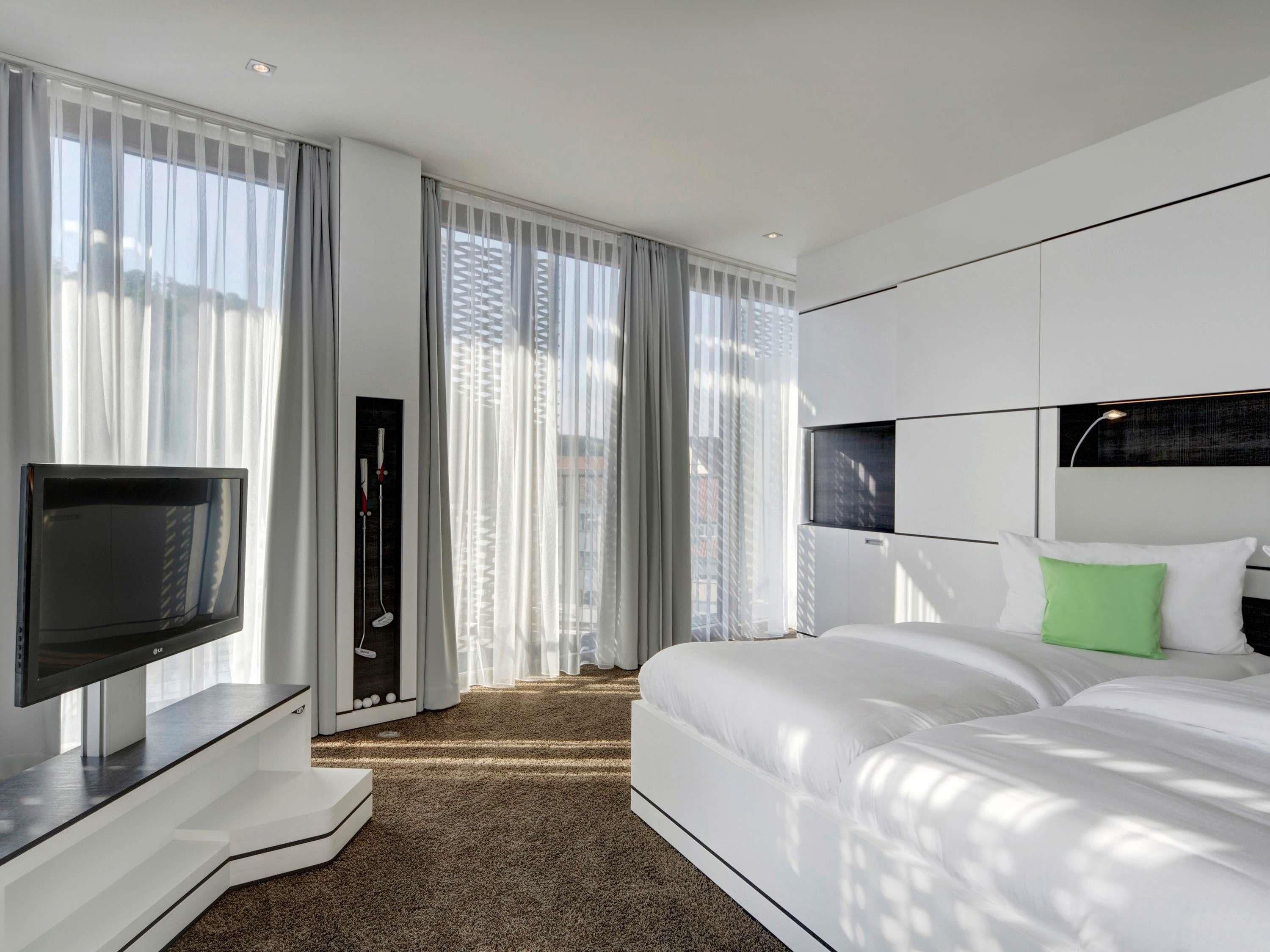 A modern hotel room with a large bed, a flat-screen TV, and a view of the city through sheer curtains.