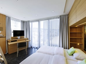 Una habitación de hotel moderna con una cama, un escritorio con televisión y amplias ventanas con cortinas.