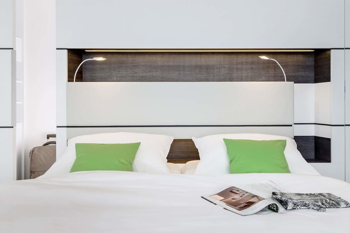 Une chambre d’hôtel moderne avec un grand lit, des oreillers verts, un magazine et une armoire visible.