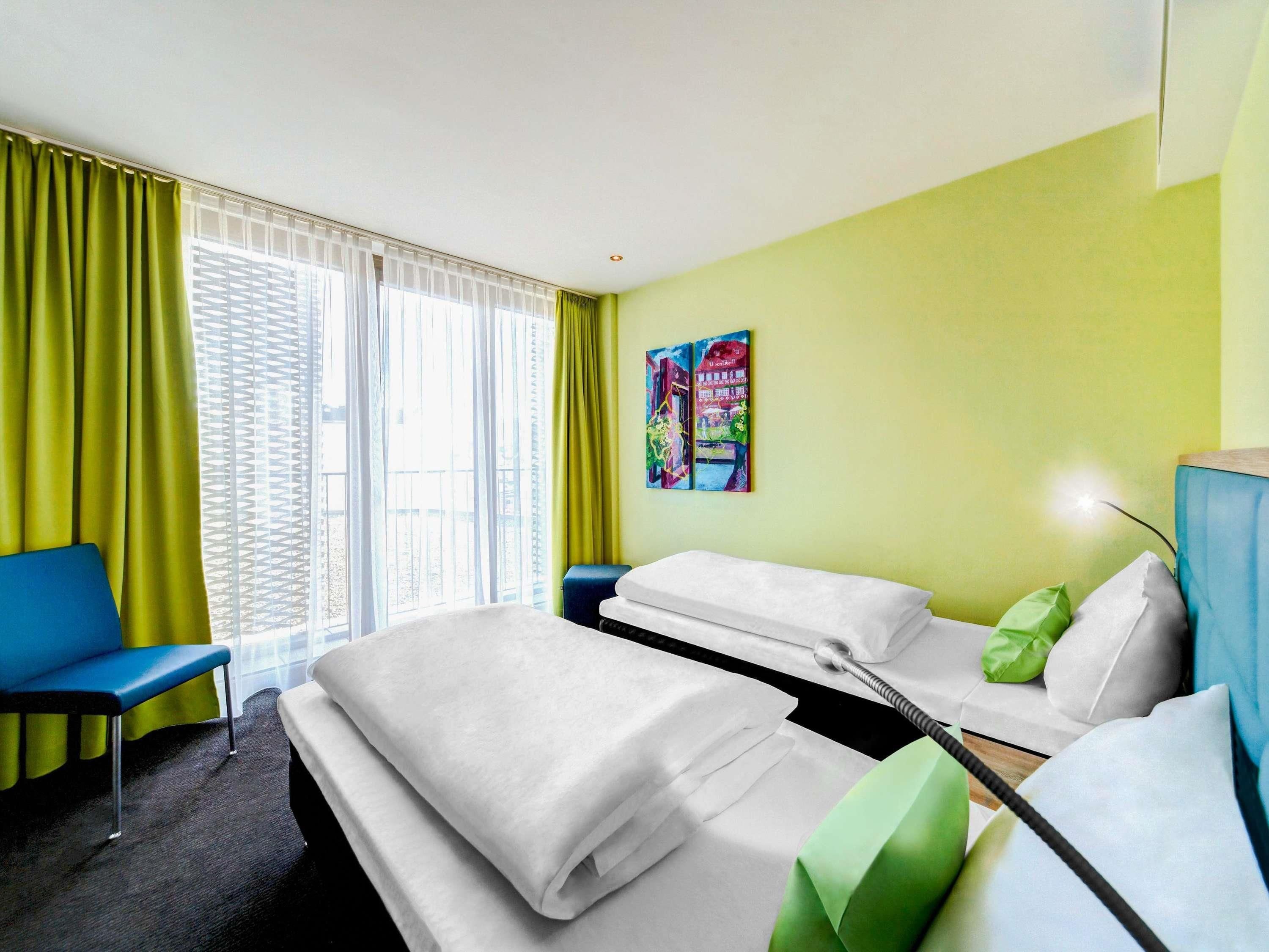 Une chambre d’hôtel avec un lit, une chaise bleue, une petite table et un tableau accroché au mur.