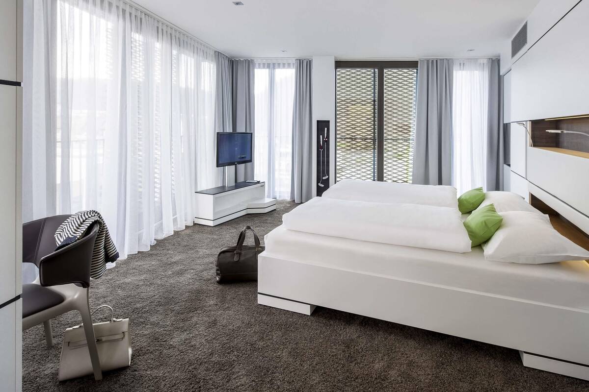 Une chambre d’hôtel moderne dotée d’un grand lit, d’un bureau avec une chaise, d’une télévision et offrant une vue sur l’extérieur grâce à de grandes fenêtres.