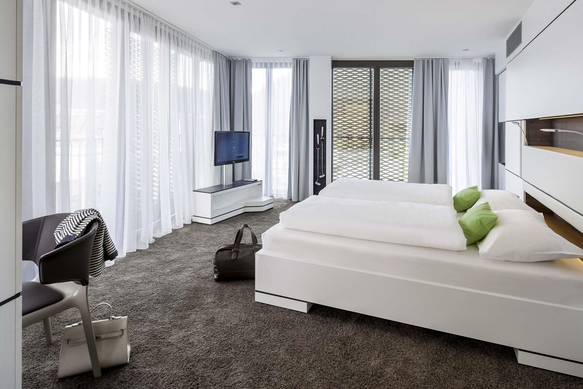 Habitación de hotel moderna con una cama grande, un escritorio con silla, televisión y vistas al exterior a través de amplios ventanales.