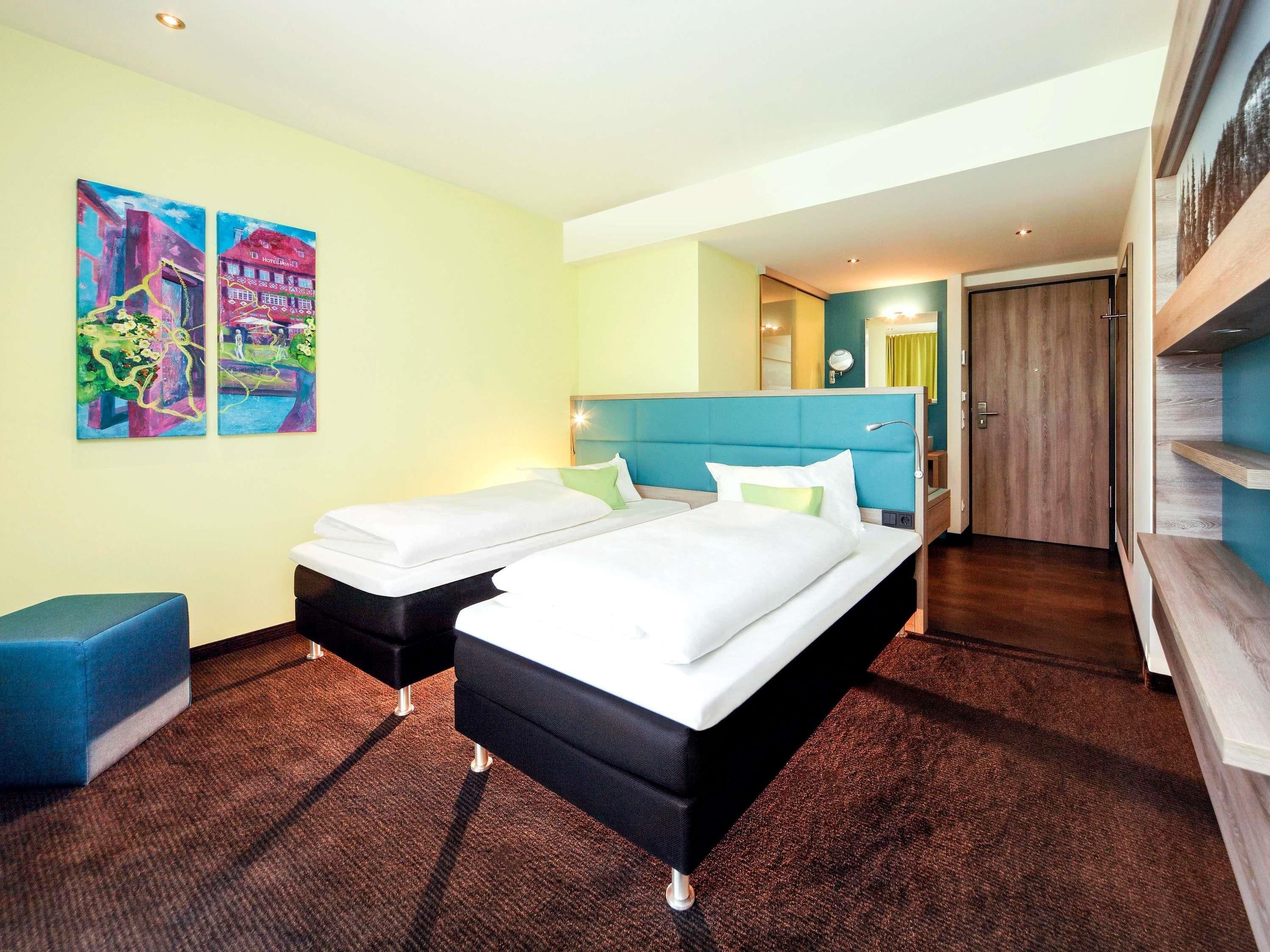 Habitación de hotel con dos camas, cabecero azul, una zona de estar pequeña y obras de arte en las paredes.
