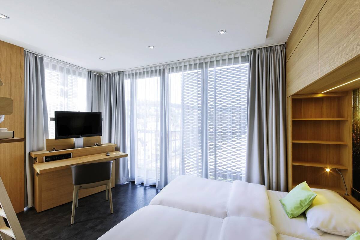 Une chambre d’hôtel moderne équipée d’un lit, d’un bureau avec une télévision et de grandes fenêtres avec des rideaux.