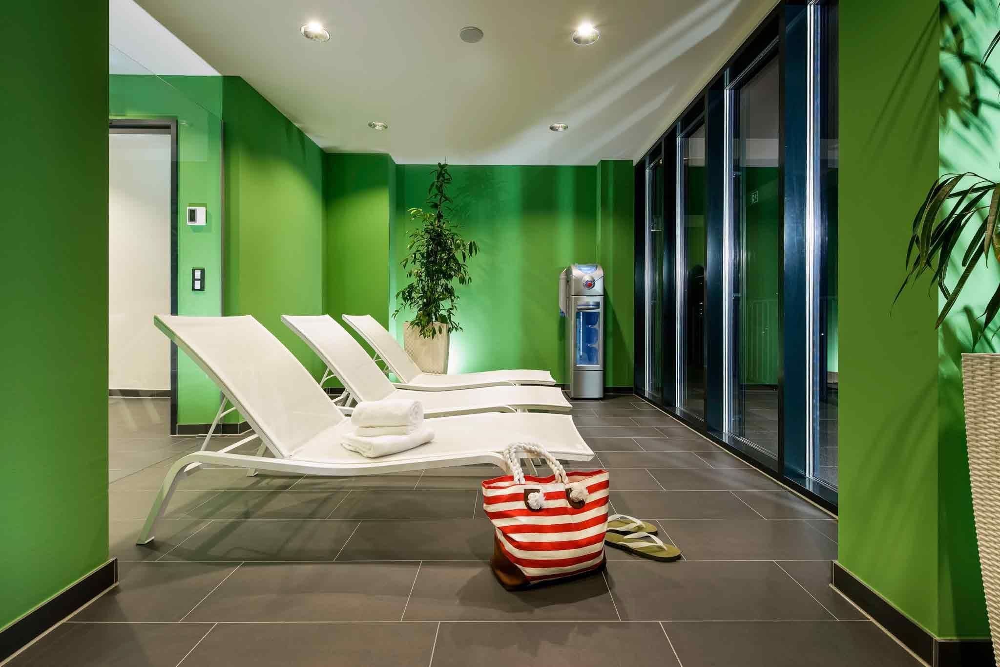 Ein moderner Wartebereich mit grünen Wänden, weißen Lounge-Sesseln, einer Kühlbox für Wasser und einem Blumentopf.