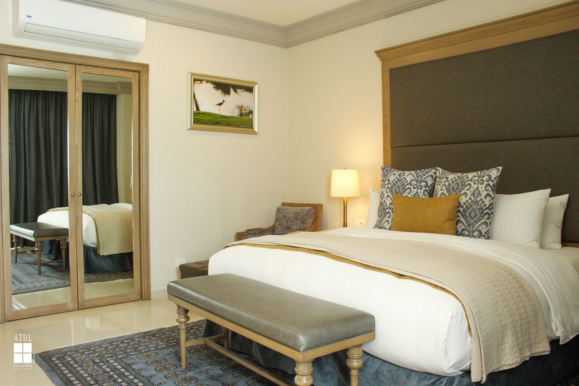 Una habitación de hotel con una cama grande, un banco, un escritorio, una silla, un espejo y un cuadro en la pared.