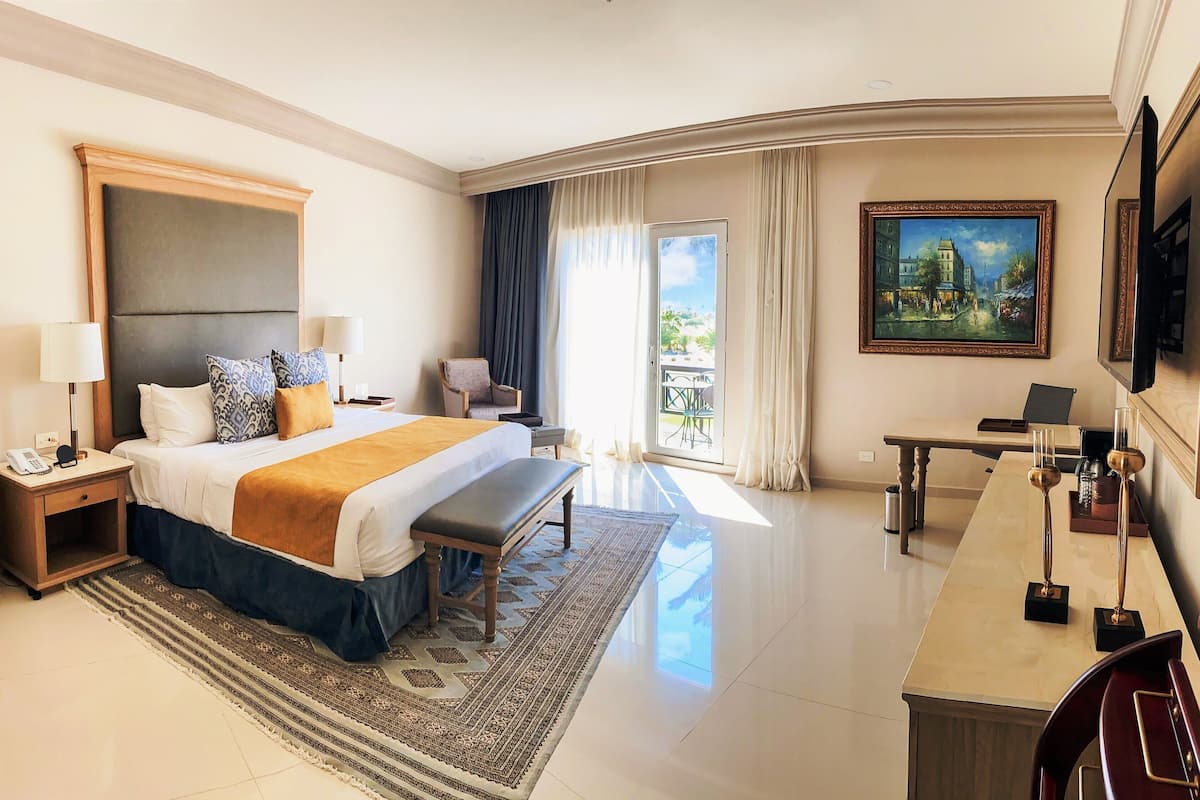 Habitación de hotel con una cama grande, mesitas de noche, un escritorio, una silla, un cuadro y vistas al exterior.