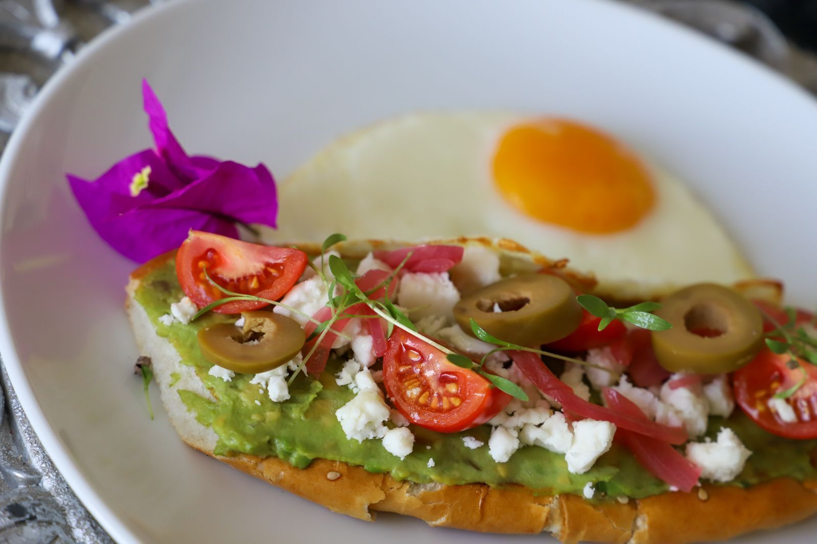 Ein Sandwich mit Avocado, Tomaten, Oliven und Feta, dazu ein Spiegelei und eine violette Blume auf einem weißen Teller.