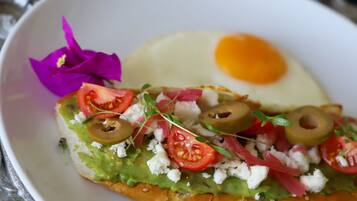 Ein Sandwich mit Avocado, Tomaten, Oliven und Feta, dazu ein Spiegelei und eine violette Blume auf einem weißen Teller.
