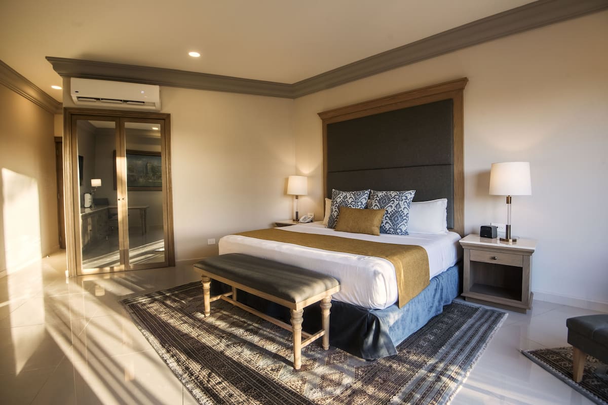 Una habitación de hotel con cama, mesitas de noche, un banco, alfombra estampada y un equipo de aire acondicionado.