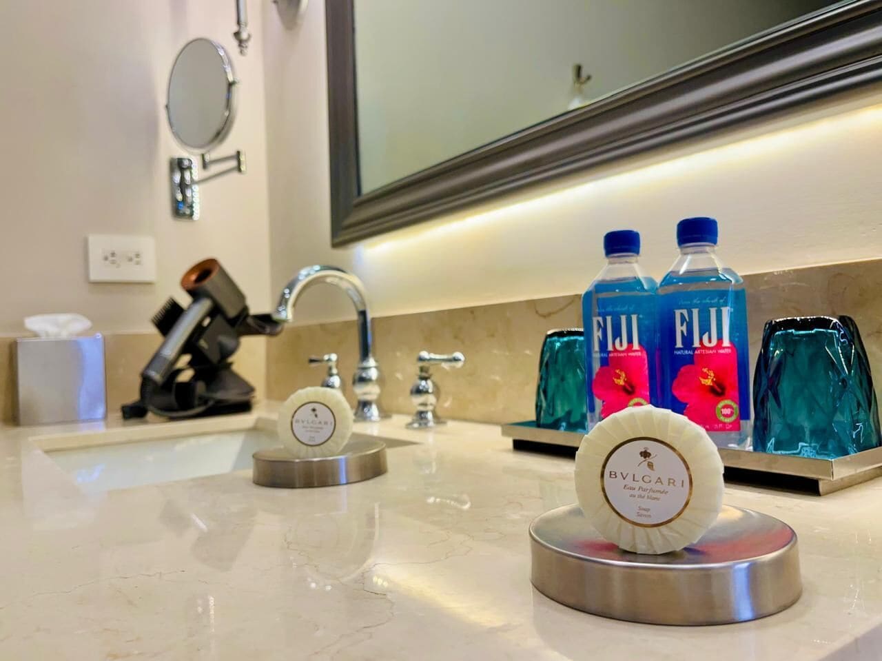 Baño de hotel con lavamanos, espejo y artículos de aseo, incluyendo agua Fiji y perfume Bulgari.