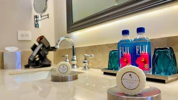 Baño de hotel con lavamanos, espejo y artículos de aseo, incluyendo agua Fiji y perfume Bulgari.