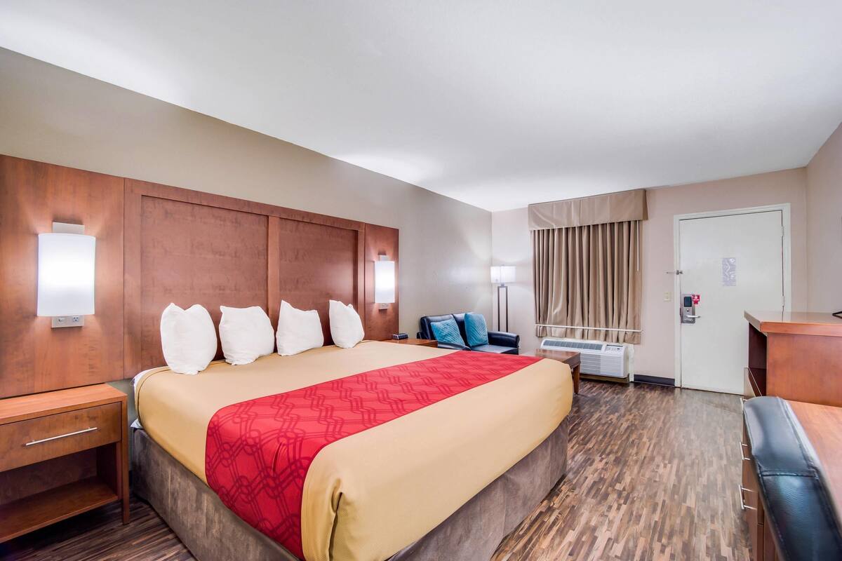 Habitación de hotel con una cama grande, mesitas de noche, un escritorio, una silla, una ventana con cortinas y un equipo de aire acondicionado instalado en la pared.