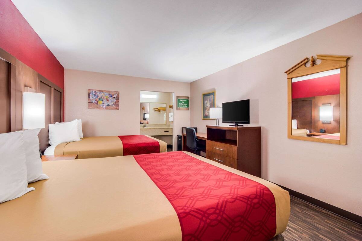 Habitación de hotel con dos camas, un escritorio, una silla, televisor, espejo y baño visible a través de una puerta abierta.