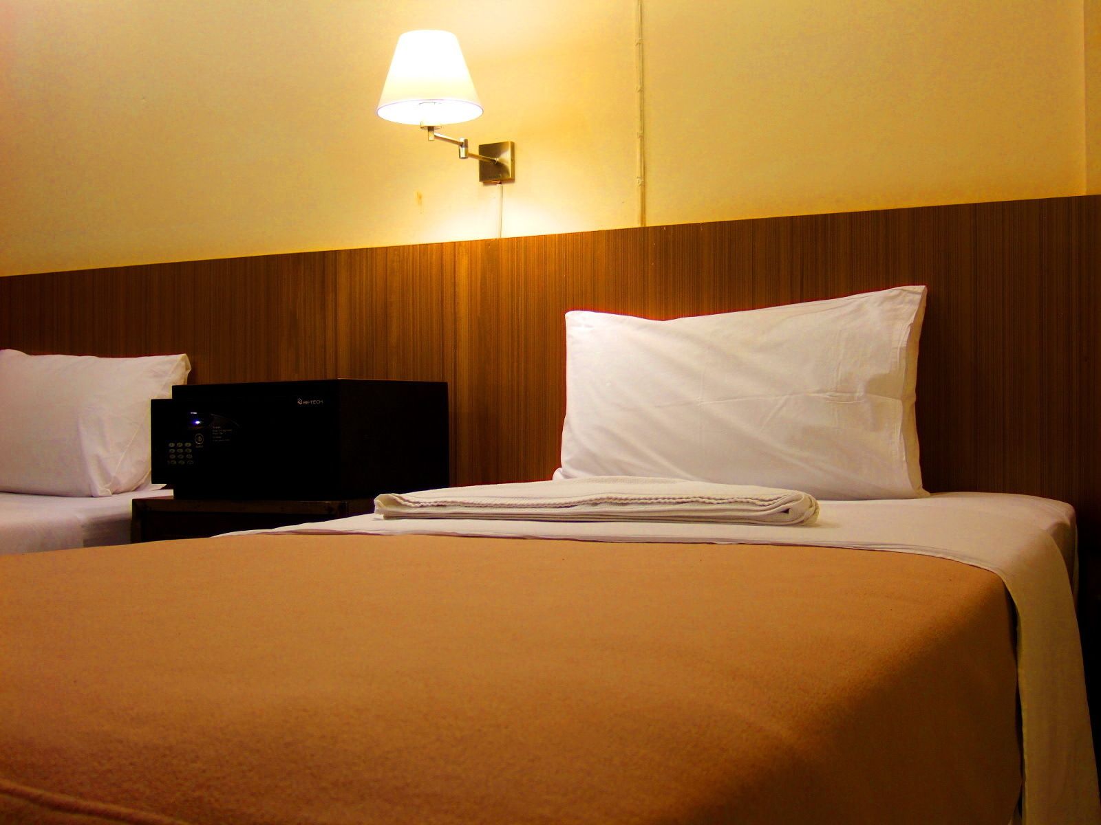 A hotel room with two beds, a bedside table with a black electronic device, a wall-mounted lamp, and a wooden panel wall.