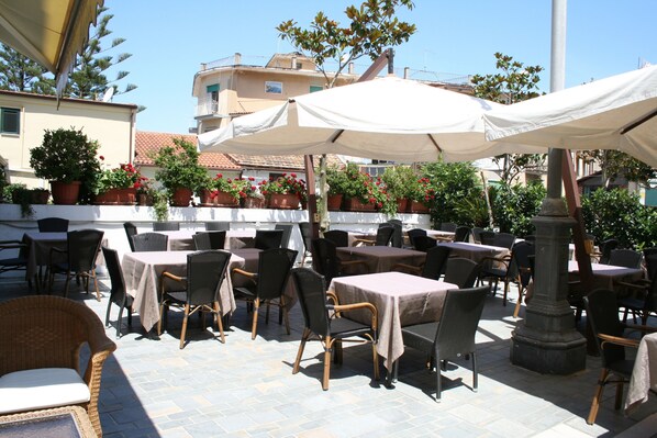 An outdoor patio with tables and chairs, surrounded by potted plants and a white canopy.