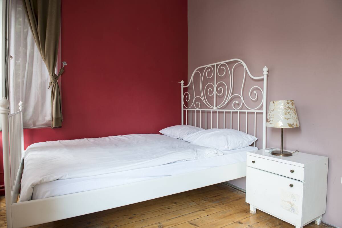 Habitación con cama individual, cabecera de metal blanco, mesita de noche con lámpara y pared roja.