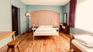 Una camera da letto con pavimento in legno, un letto con testiera bianca, un comodino, una lampada, una sedia e una finestra con tende.