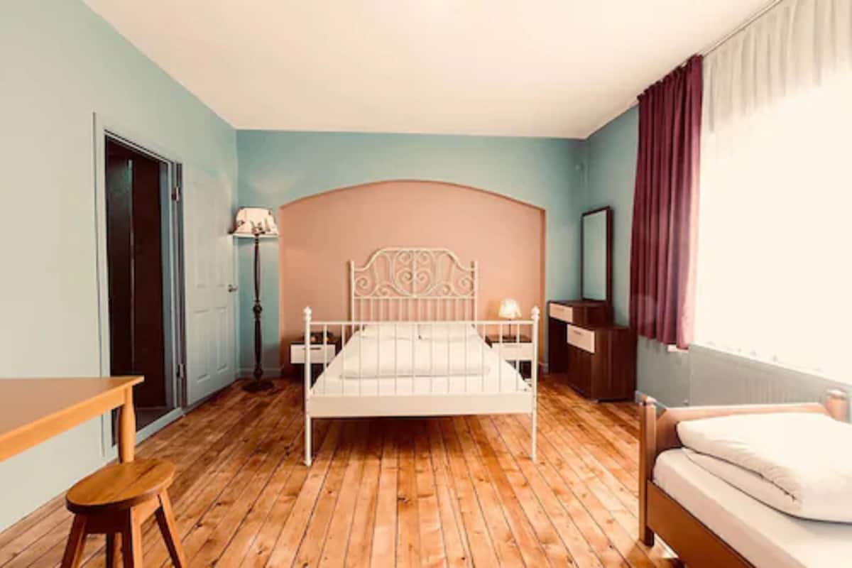 Un dormitorio con piso de madera, una cama con cabecera blanca, una mesita de noche, una lámpara, una silla y una ventana con cortinas.