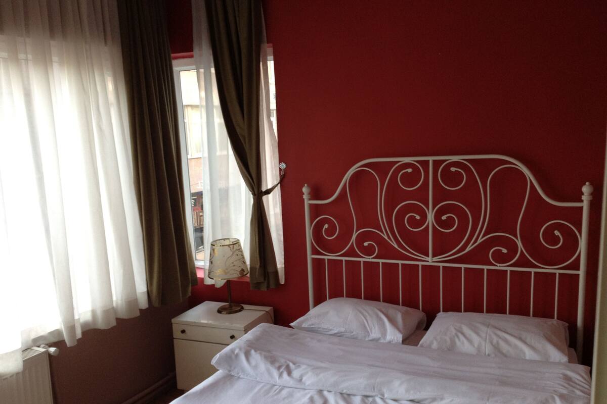 Un dormitorio con cabecera de metal, ropa de cama blanca, una mesita de noche con lámpara y una ventana con cortinas.