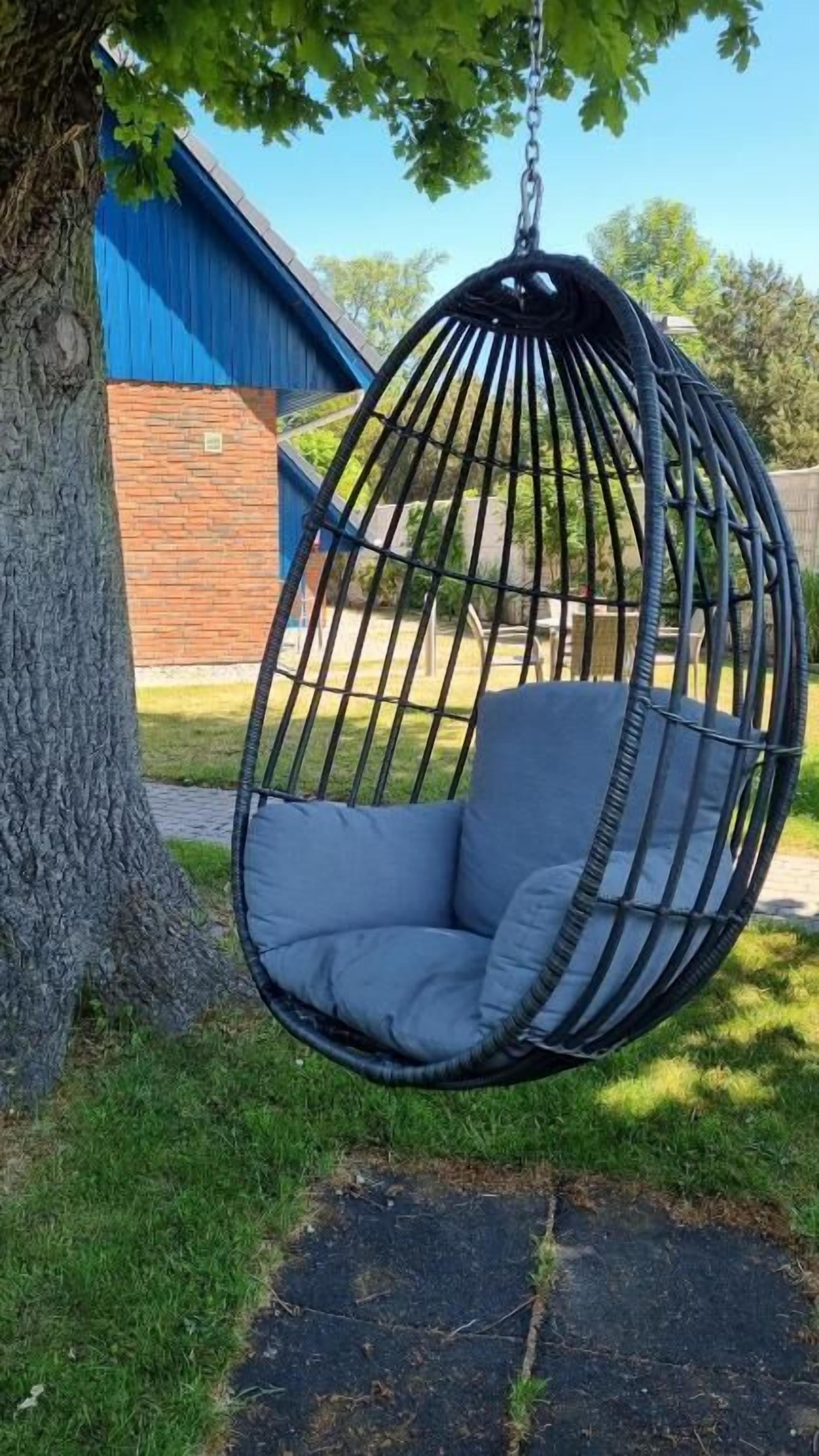 A black hanging chair with a cushion, attached to a tree, in a backyard with a blue building in the background.